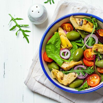 Green salad with artichokes, tomatoes and olives in white bowl.